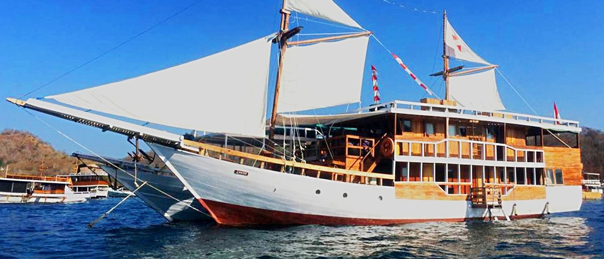 A large wooden boat with white sails floats on blue water under a clear sky, with hills in the background.