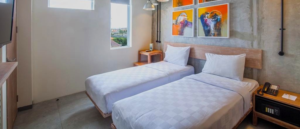 A bright hotel room contains two single beds, white bedding, and colourful abstract artwork on the wall.