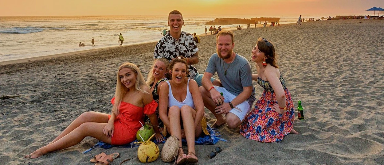 Six young people sit on a sandy beach with coconuts as the sun sets over the ocean.