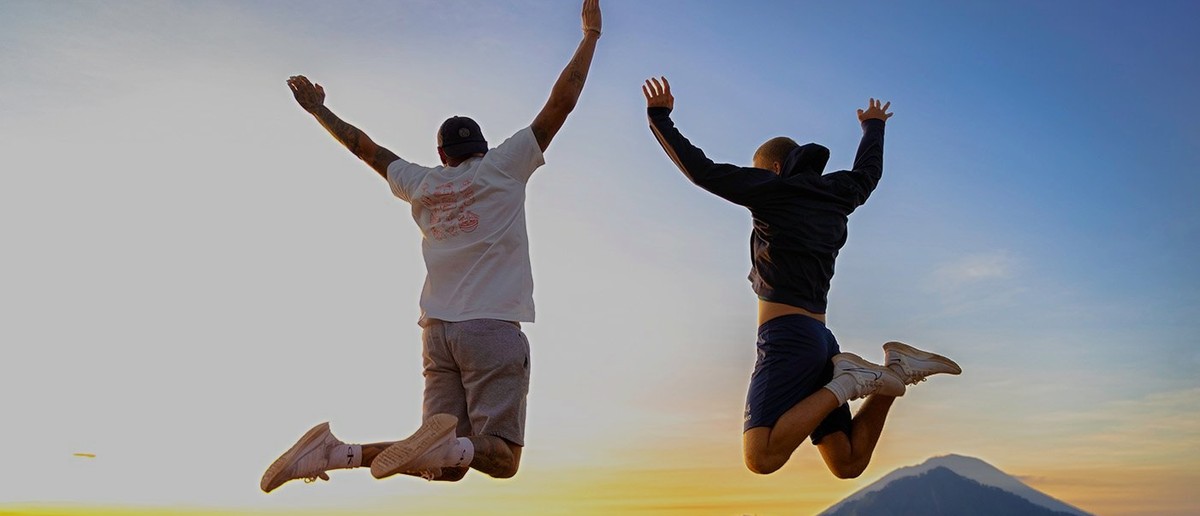 Two people jump with raised arms against a colourful sunrise sky above mountain peaks.