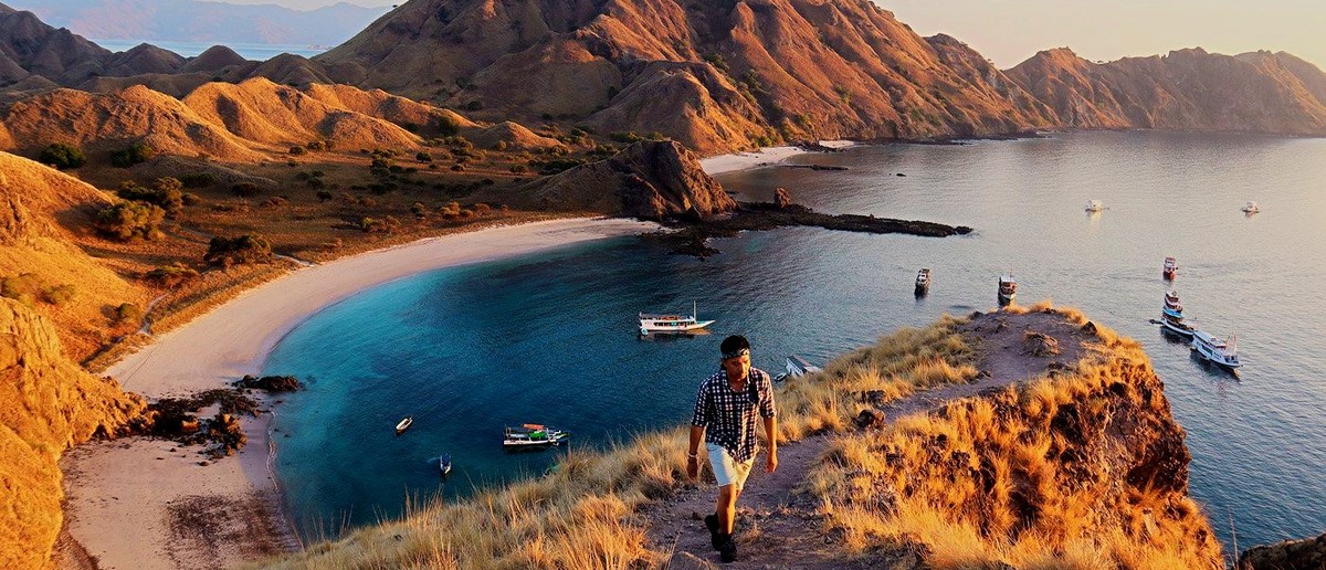 A person walks a dry grassy path above a bay with turquoise water, white sand, and distant mountains.