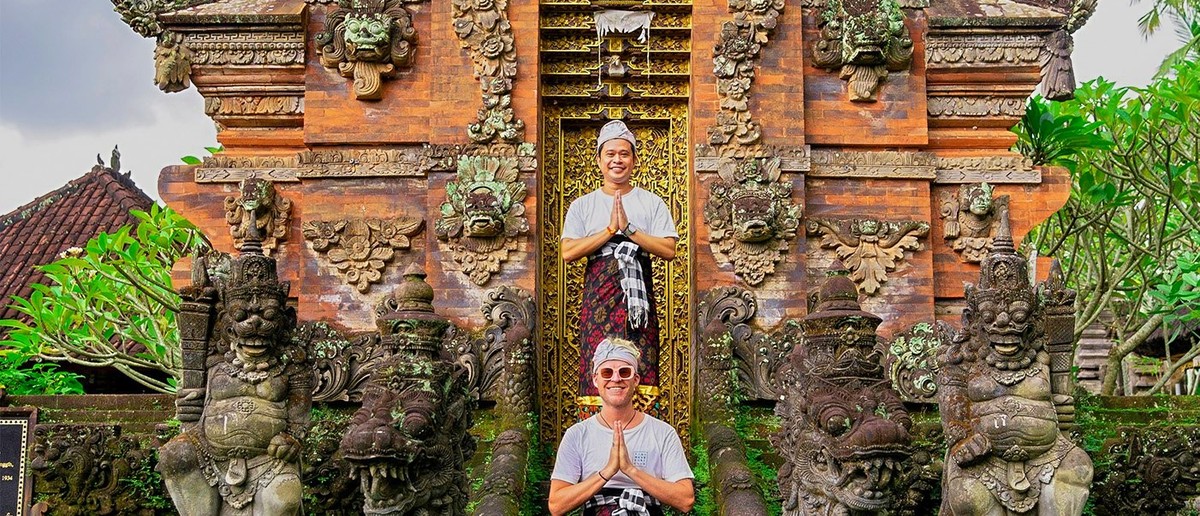 Two men in traditional dress pose with hands pressed together before an ornate brick temple gate.