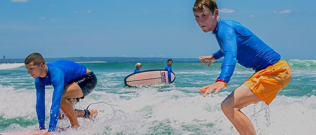 Two young men in blue tops are riding surfboards on green ocean waves.