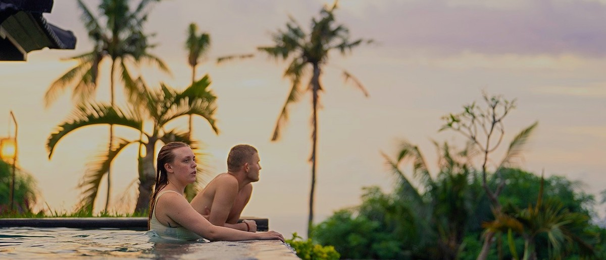 Two people relax in an infinity pool, looking out at palm trees against a sunset sky.