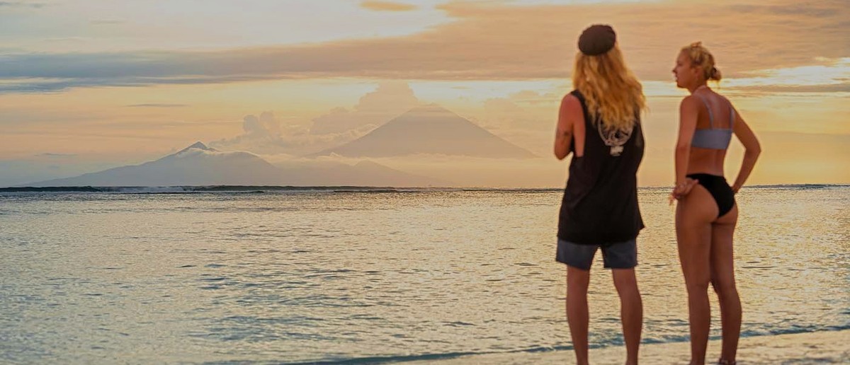 Two people stand in the shallow sea, looking at distant mountains under a colourful sunset sky.