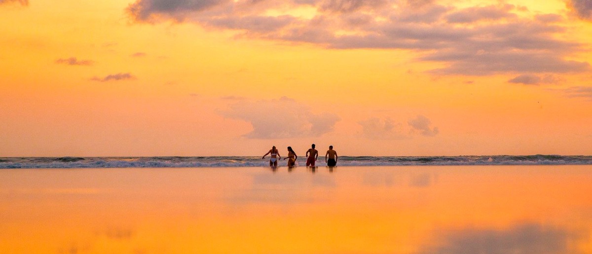Four people wade in the ocean at sunset, with the sky and water bathed in orange and yellow hues.