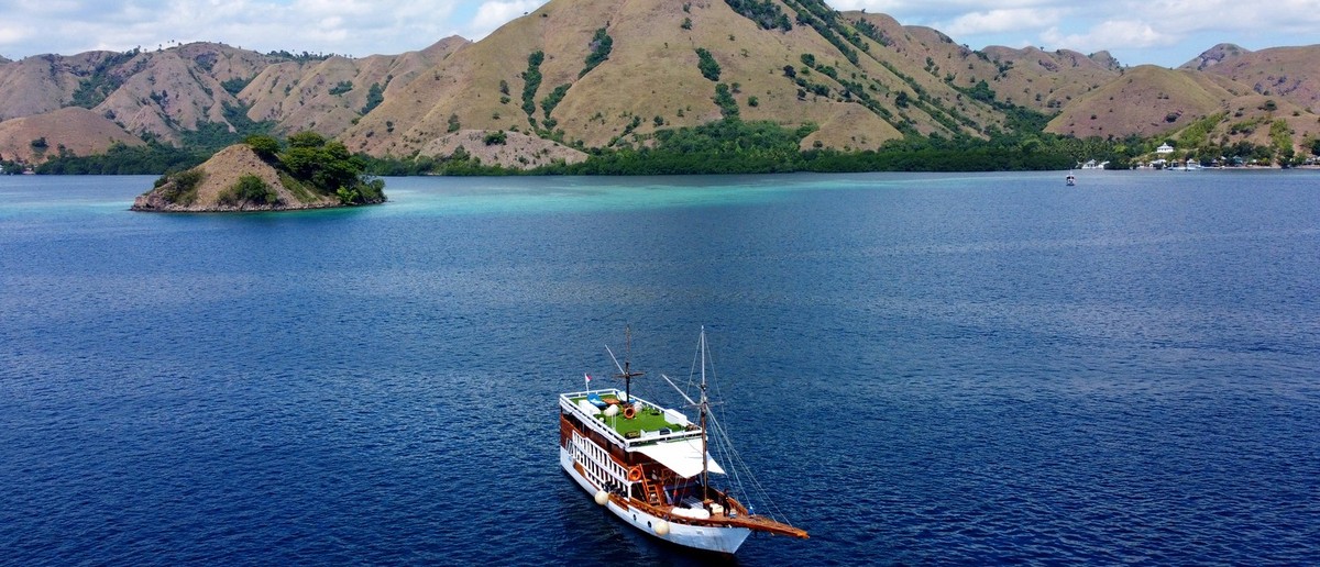 A large wooden sailing boat glides across deep blue water with dry, hilly islands in the background.