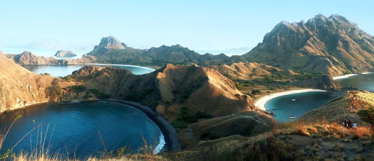 Rolling dry hills surround deep blue bays with white and black sand beaches under a clear sky.