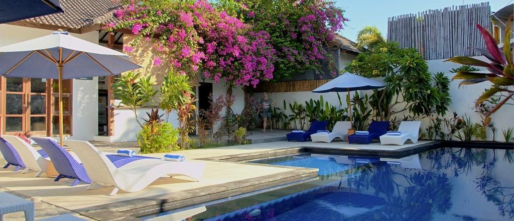 A sunny swimming pool area with sun loungers, blue umbrellas, and a building covered in vibrant pink flowers.