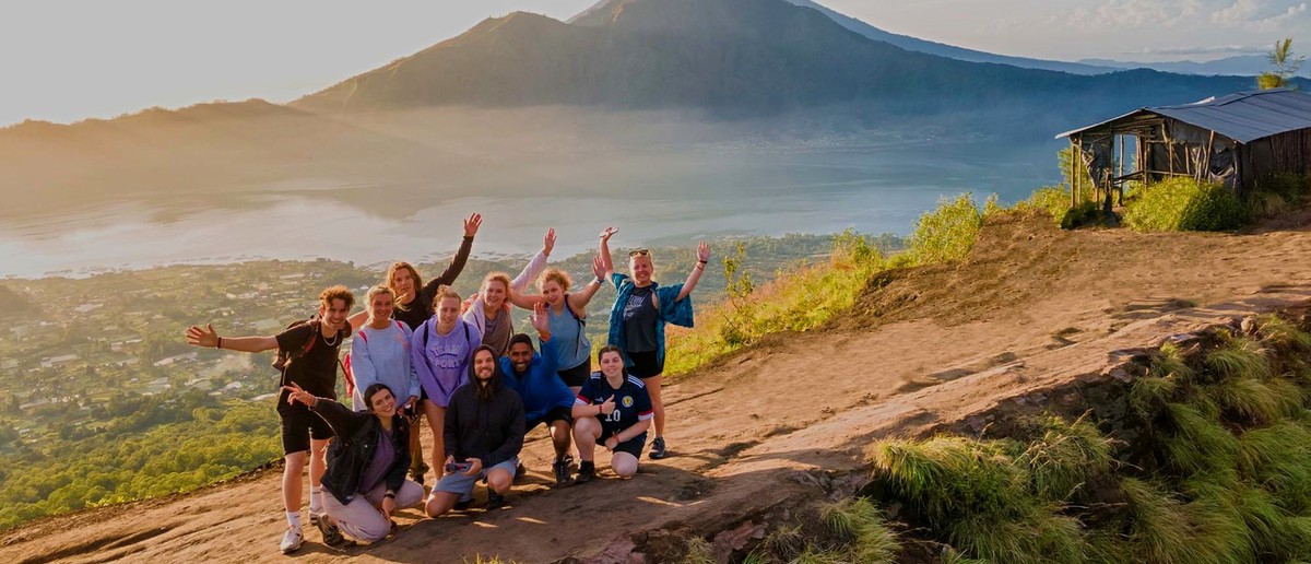 A group of people pose on a mountain slope at sunrise, overlooking a misty lake and distant volcanoes.