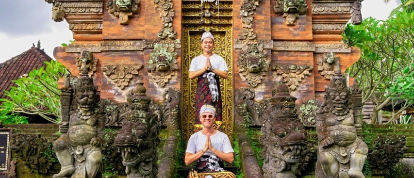 Three travellers in traditional sarongs pose with hands together on moss-covered temple steps.