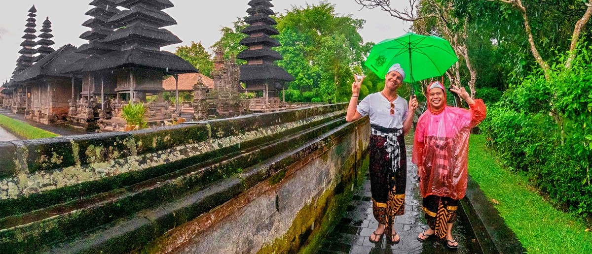 Two people pose with peace signs on a wet path, with multi-tiered temple structures and lush greenery behind them.