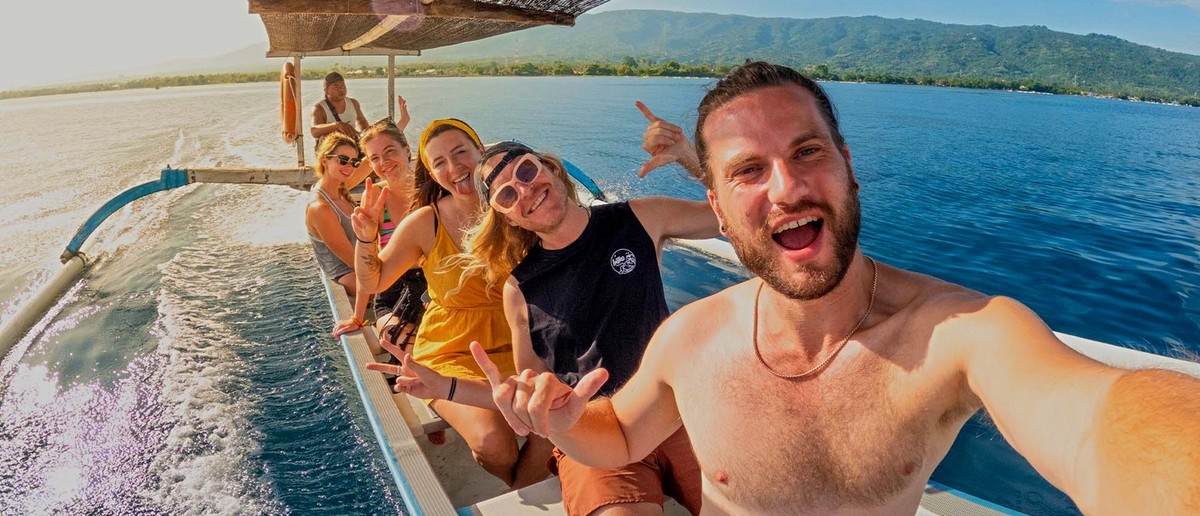 Six smiling people on an outrigger boat on bright blue water with distant hills.