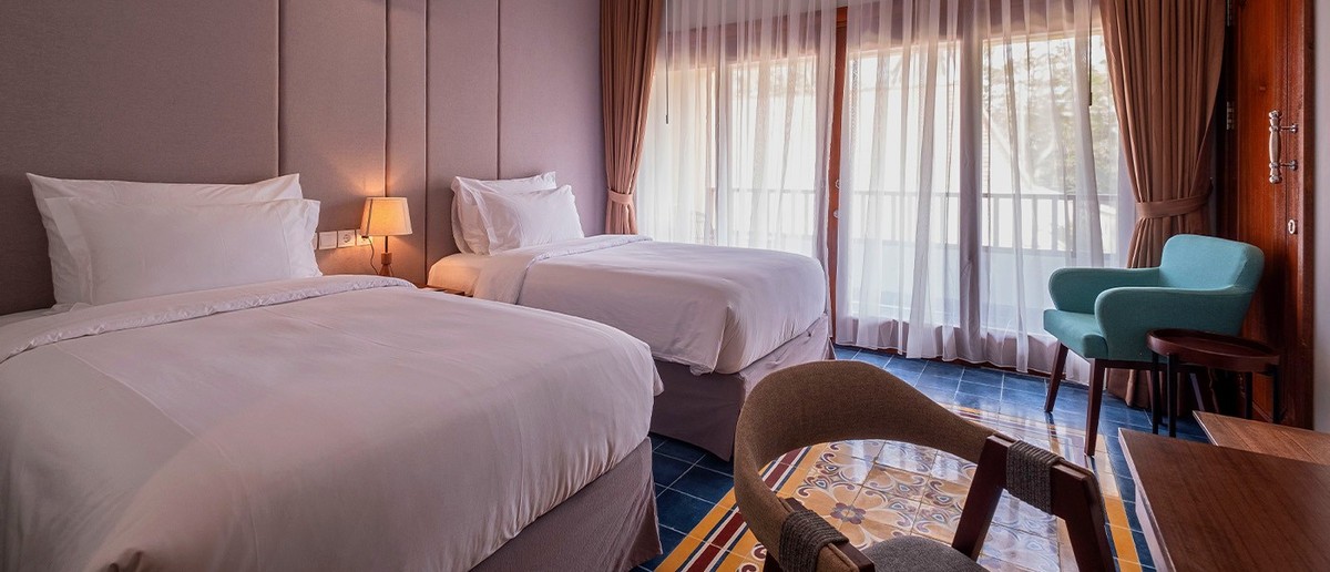 Hotel room with two single beds, a teal armchair, and patterned floor tiles next to a window.