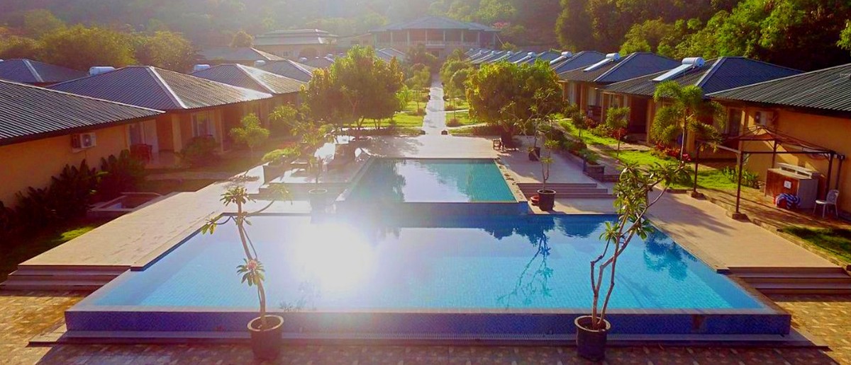 A large swimming pool reflecting sunlight, surrounded by rows of bungalows, with lush green hills in the background.