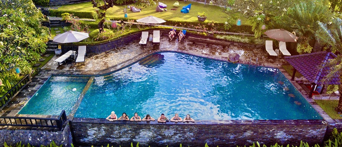 A private villa with a large swimming pool, people relaxing in the water, surrounded by lush gardens and palm trees.