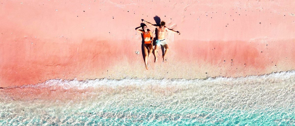 Two people lie on a pink sand beach beside clear turquoise water with gentle waves.