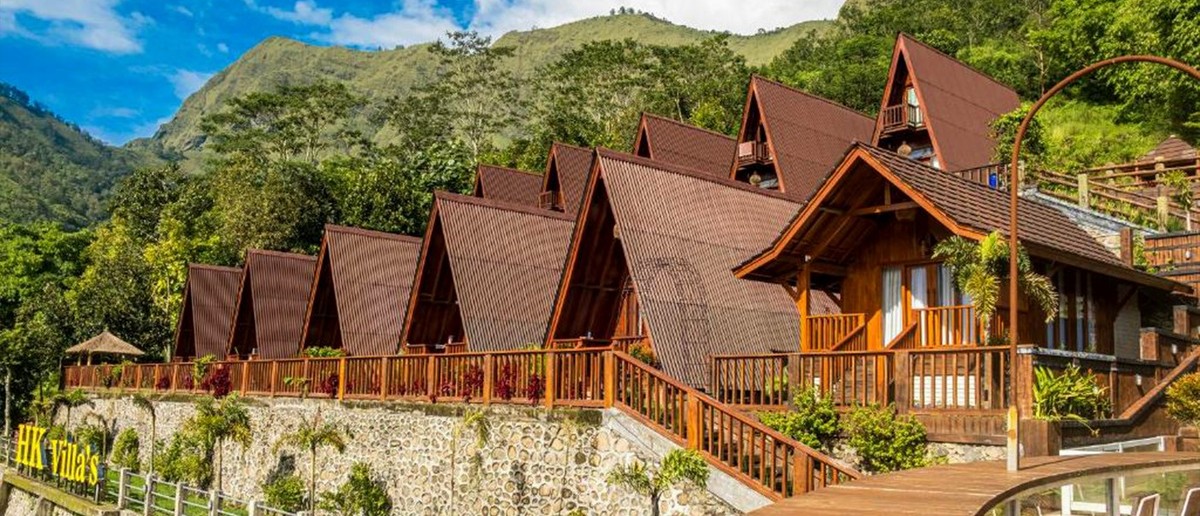 Several wooden A-frame villas with brown roofs are built into a lush green mountain hillside.