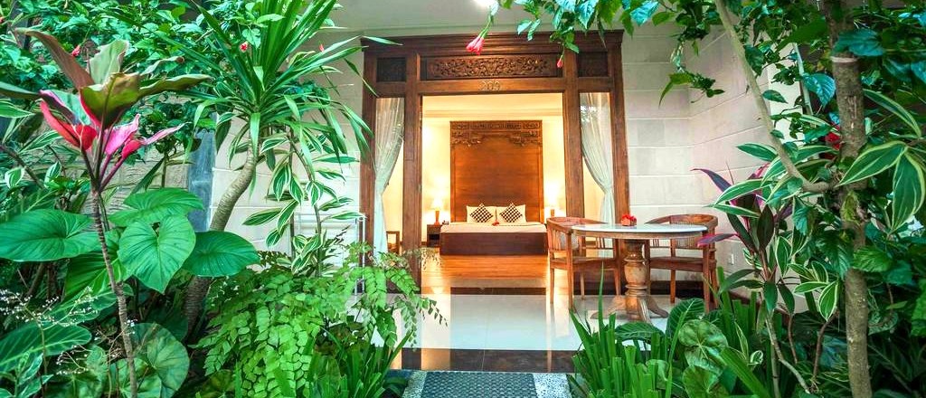A wooden doorway leads to a bright bedroom with a bed, surrounded by lush tropical foliage.