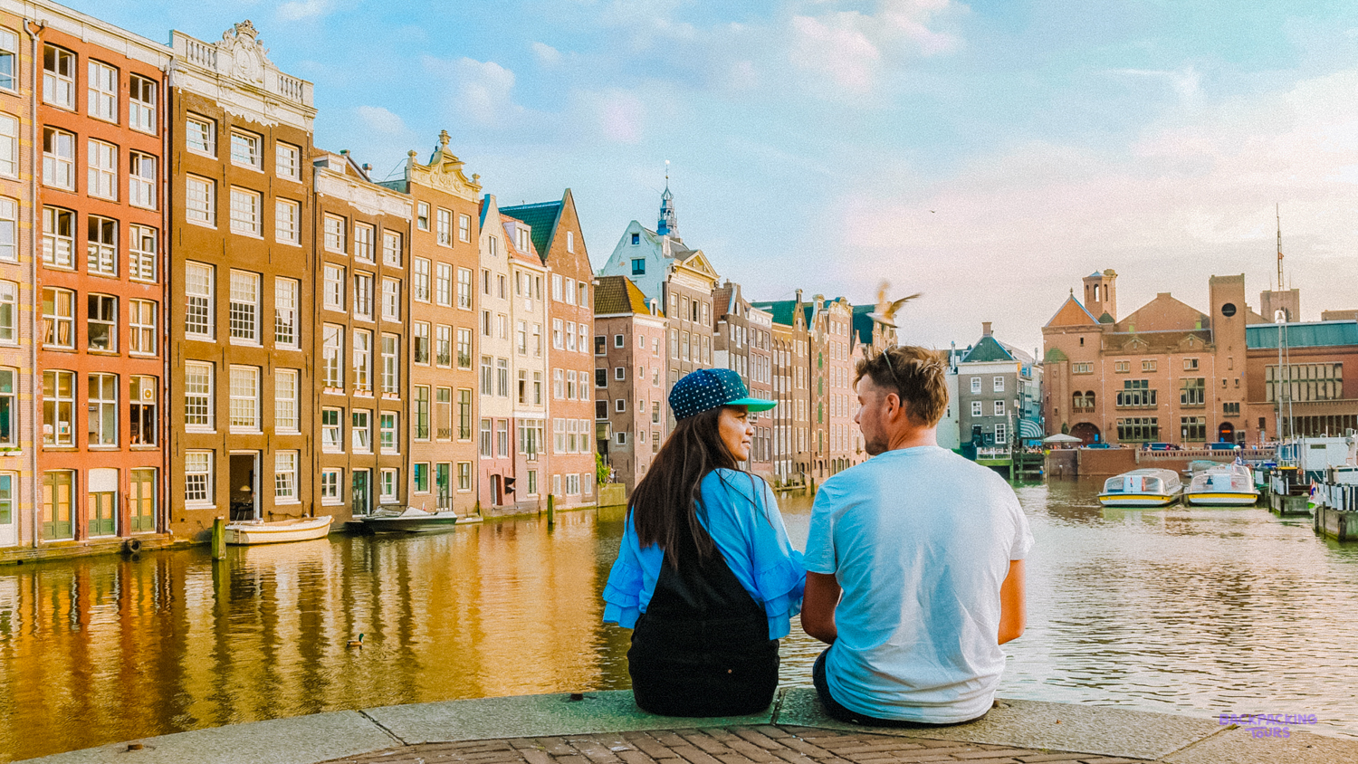 Two travellers sit by an Amsterdam canal lined with tall, narrow brick houses.