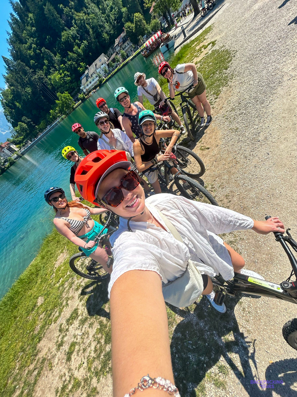 A group of cyclists in helmets takes a selfie beside turquoise water near Bled.