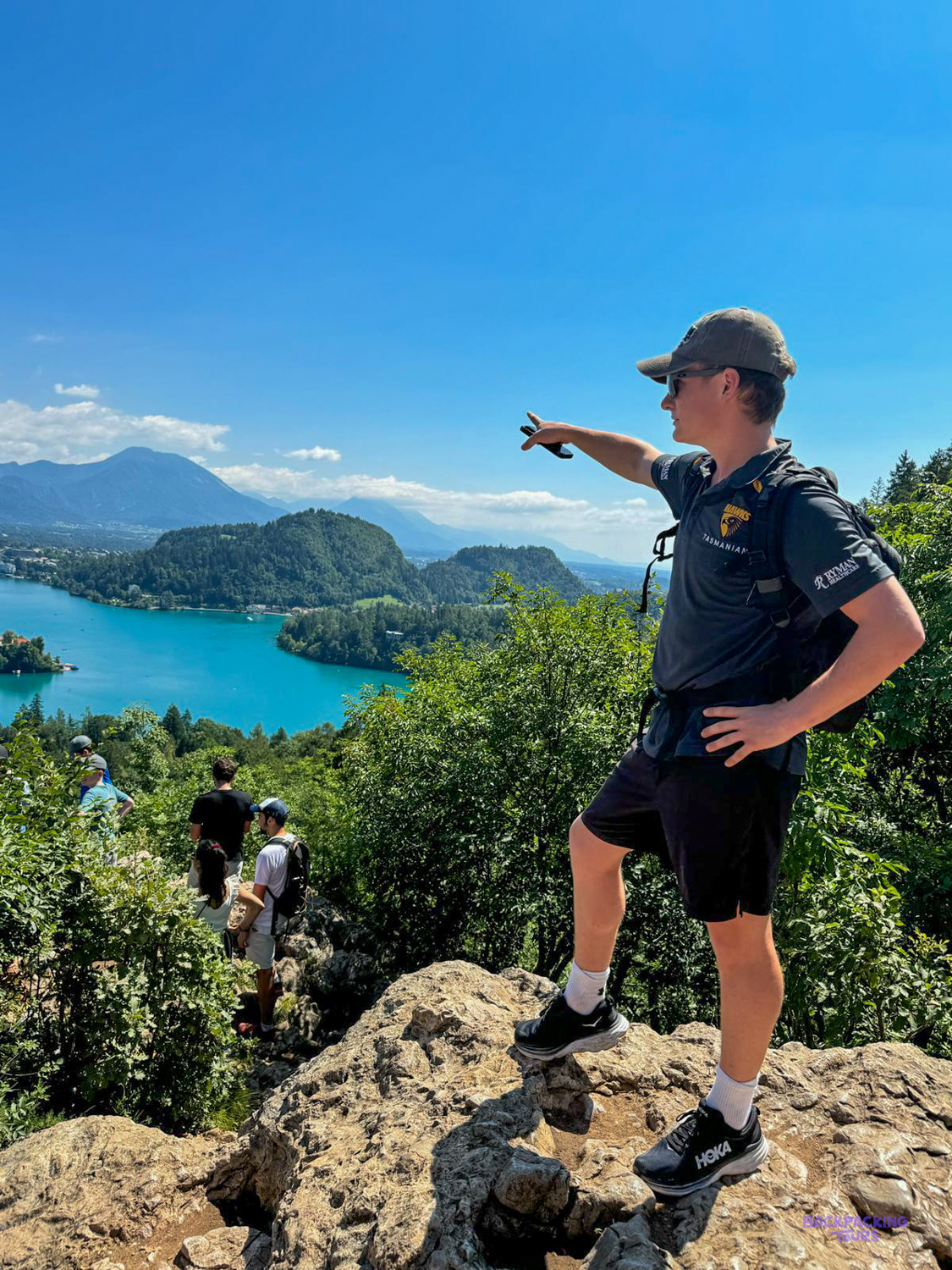 A man on a rocky viewpoint points out over the turquoise water of Lake Bled.