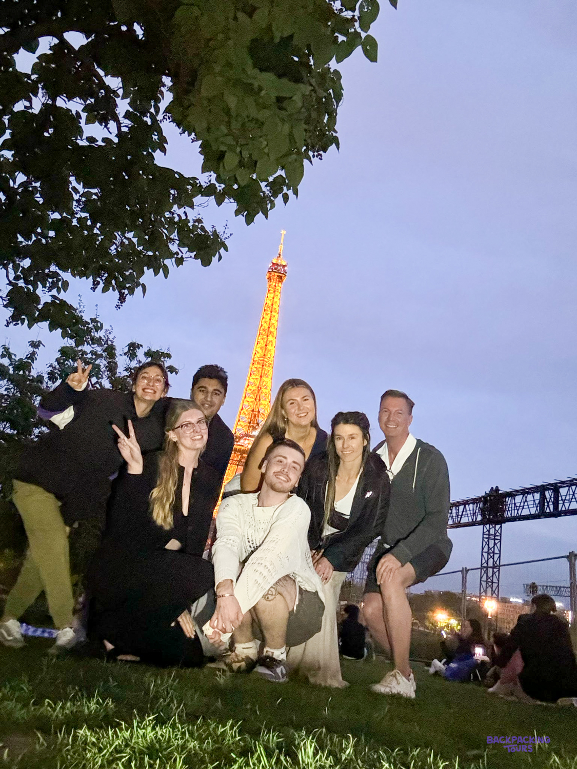 A group of travellers poses together at dusk with the lit-up Eiffel Tower behind them.