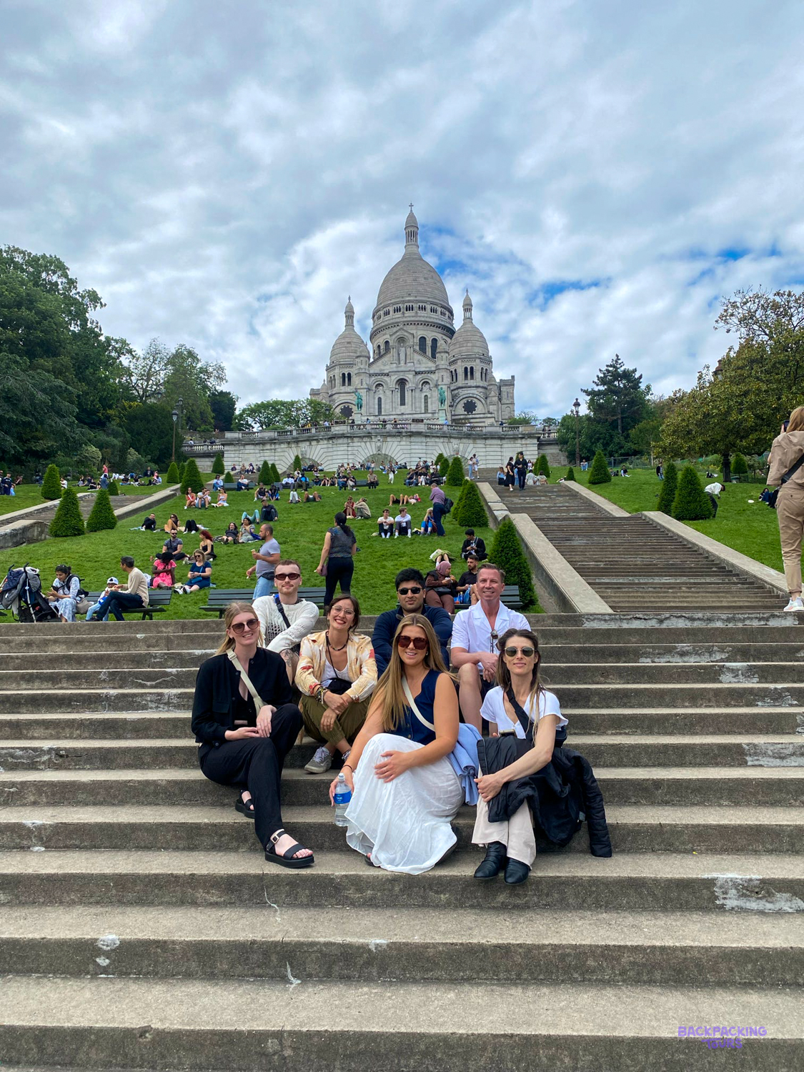 Travellers sit together on the steps below the white domes of the Sacré-Cœur in Montmartre.