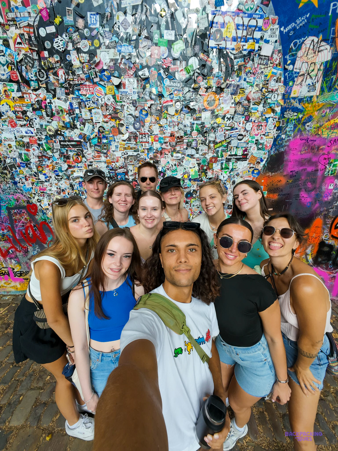 A group of travellers takes a selfie in front of a wall covered in graffiti and stickers.