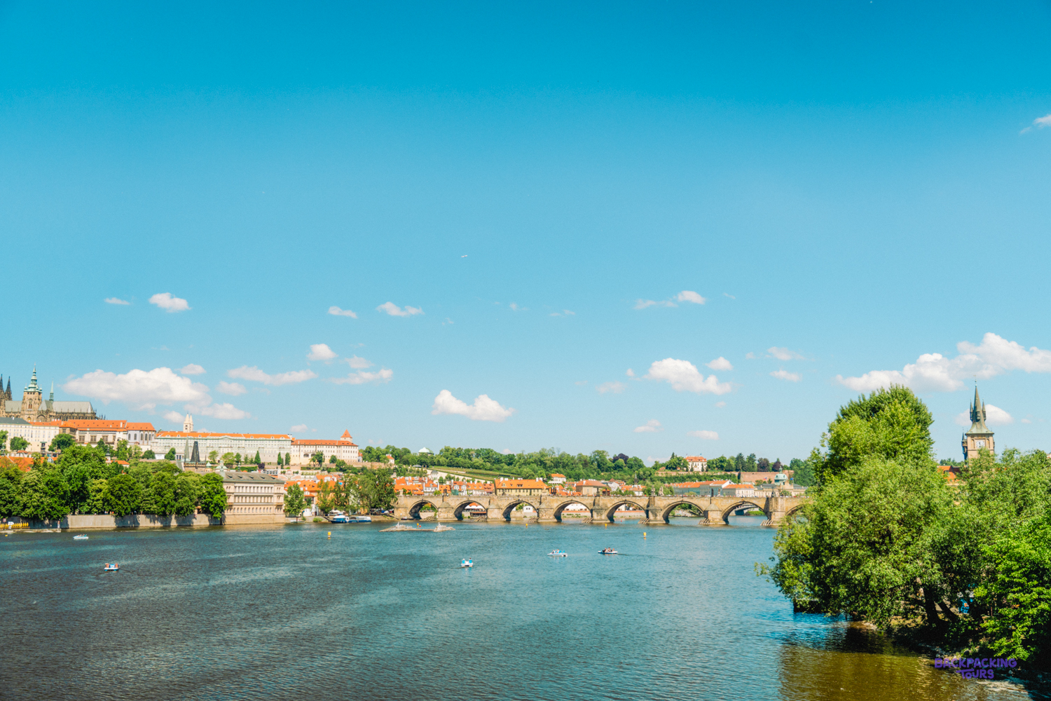 The wide Vltava river in Prague on a sunny day, with a long stone bridge across it.