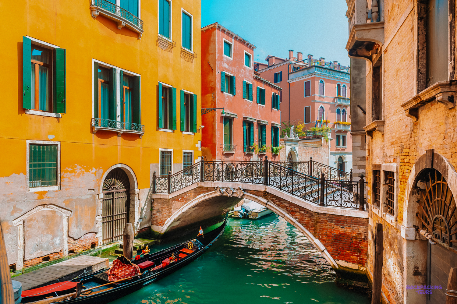A gondola passes under an iron bridge on a Venice canal lined with orange and red houses.