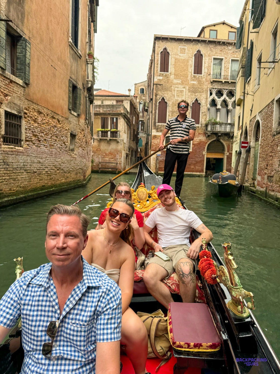 Travellers smile from a gondola as the gondolier steers them along a narrow Venice canal.