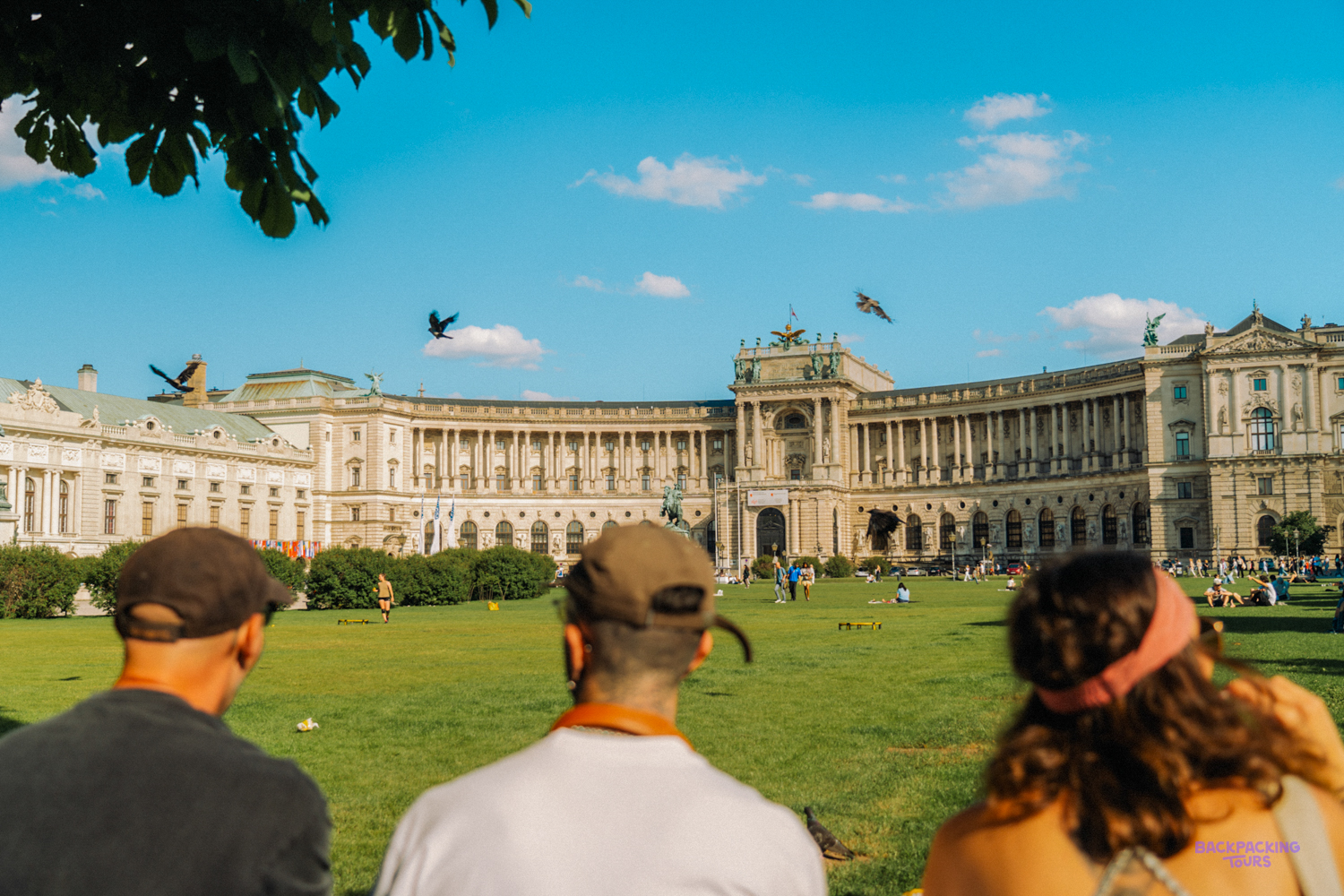 Travellers look across a sunny lawn towards the curved front of the Hofburg Palace in Vienna.
