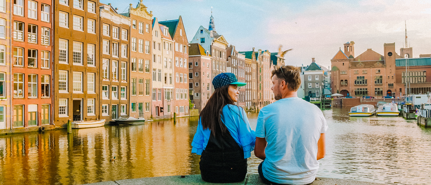 Two travellers sit by an Amsterdam canal lined with tall, narrow brick houses.