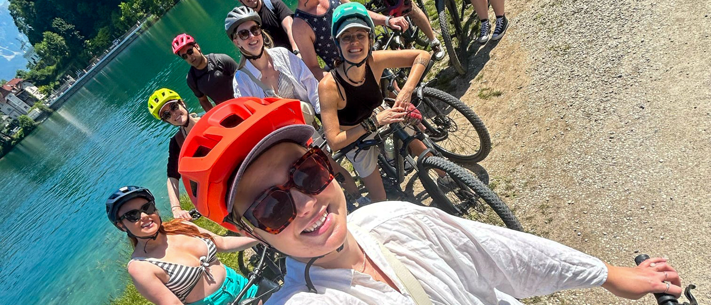 A group of cyclists in helmets takes a selfie beside turquoise water near Bled.