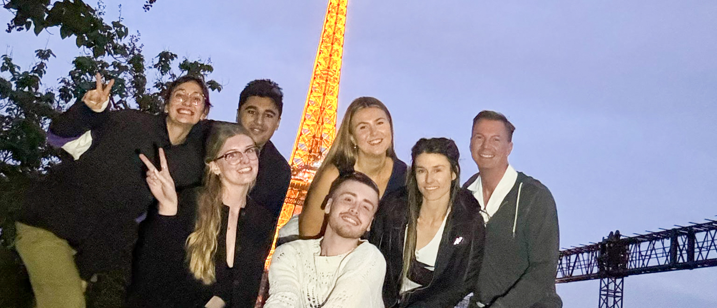 A group of travellers poses together at dusk with the lit-up Eiffel Tower behind them.