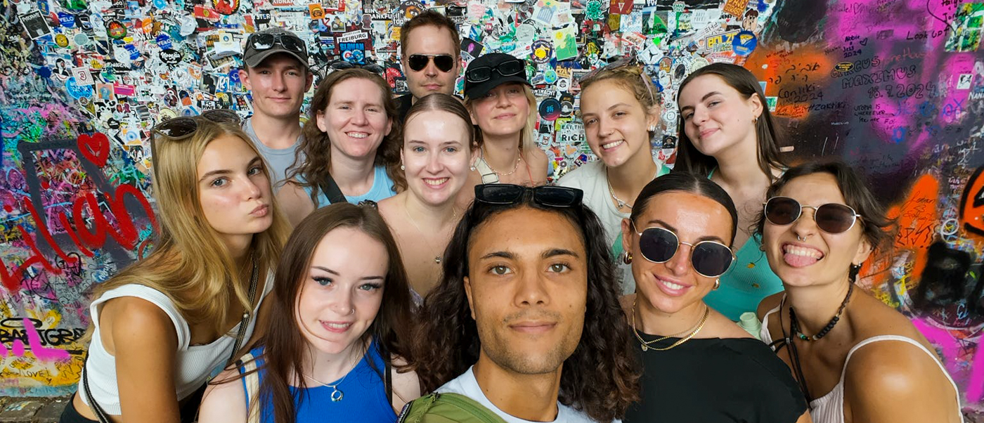 A group of travellers takes a selfie in front of a wall covered in graffiti and stickers.