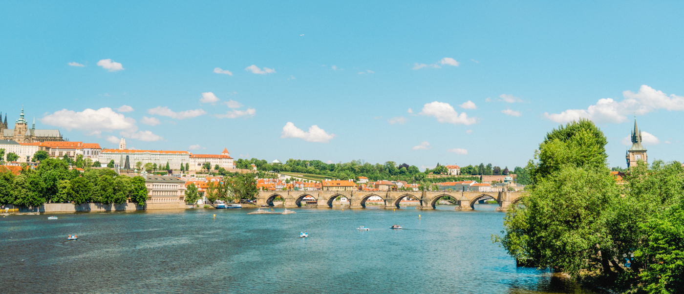 The wide Vltava river in Prague on a sunny day, with a long stone bridge across it.