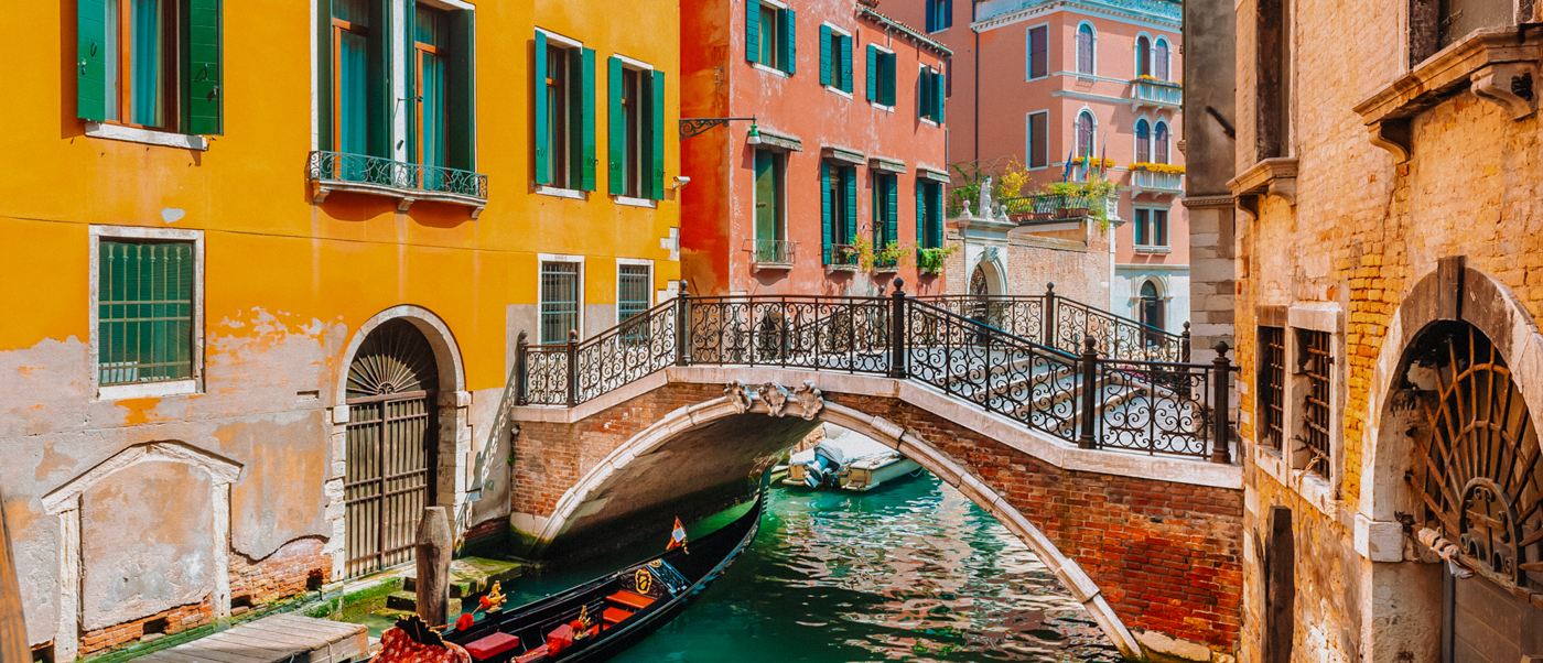 A gondola passes under an iron bridge on a Venice canal lined with orange and red houses.