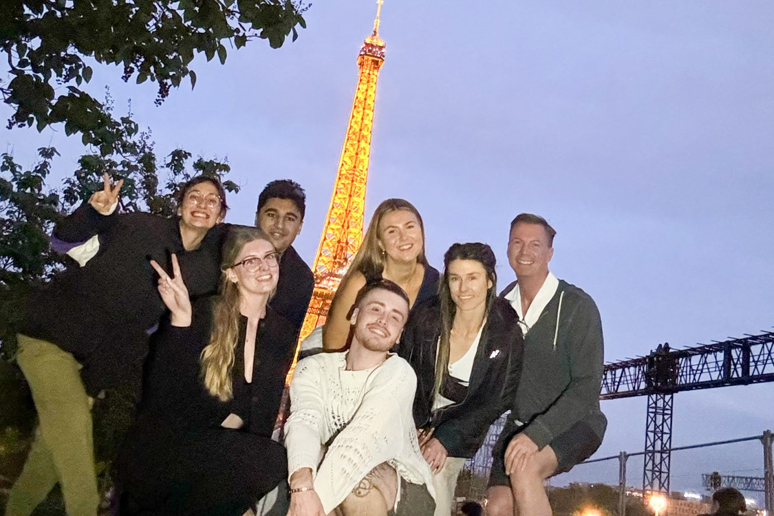A group of travellers poses together at dusk with the lit-up Eiffel Tower behind them.
