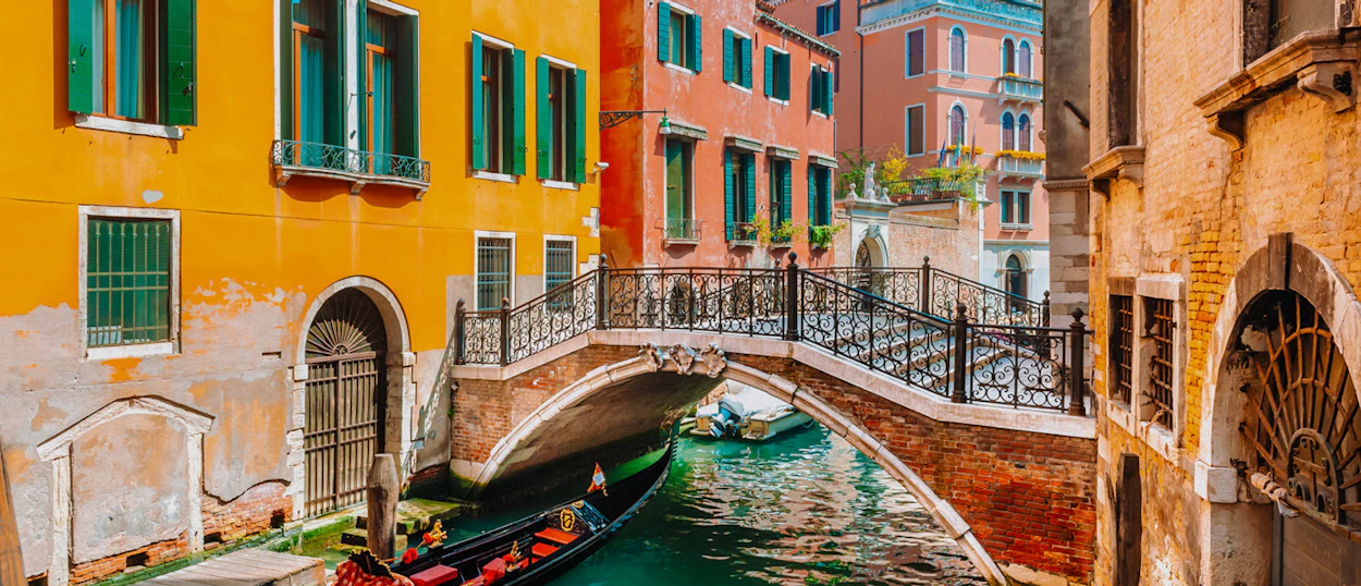 A traditional gondola glides under a stone bridge between vibrant, historic buildings in Venice.