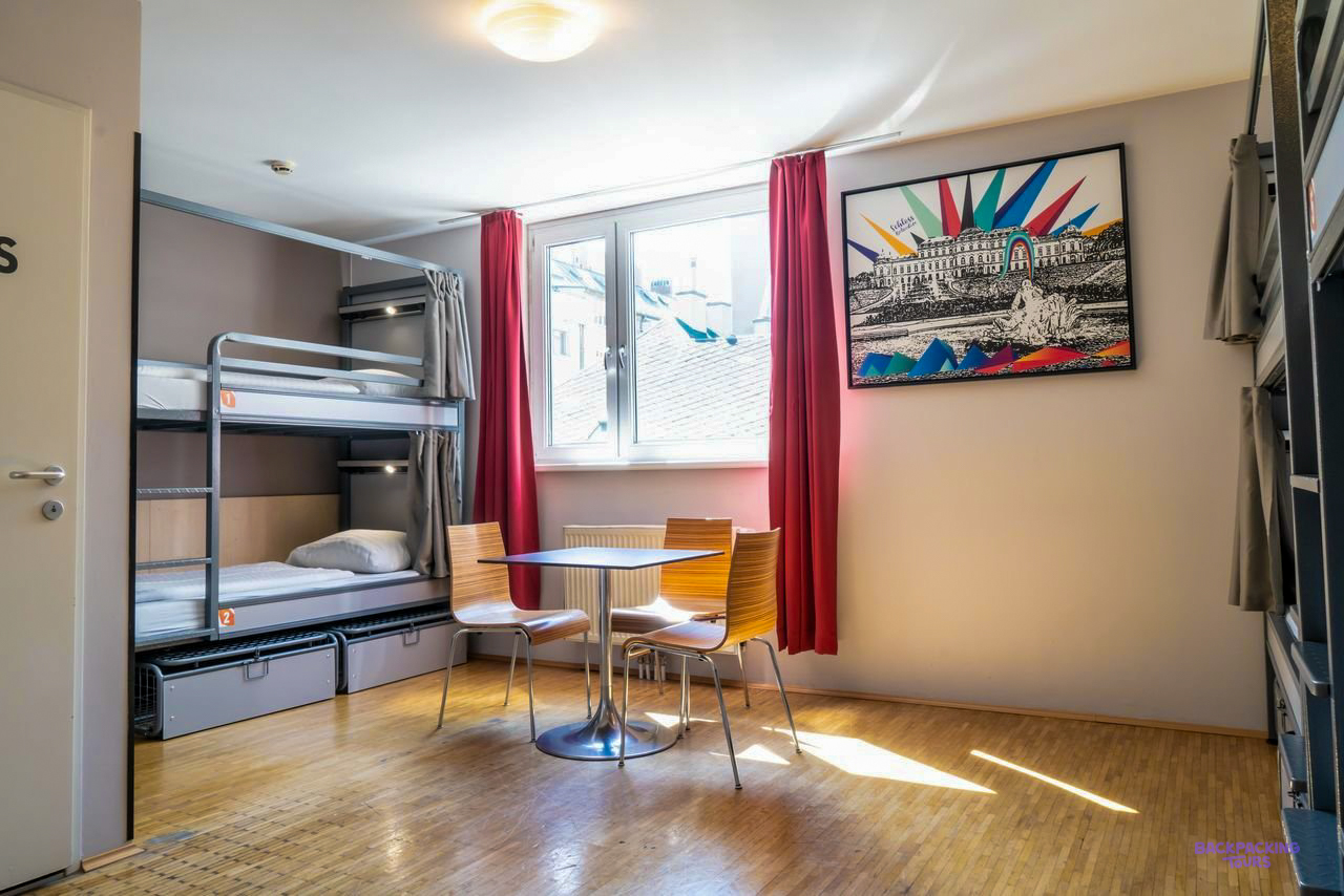 A dorm room with metal bunk beds, red curtains and a small table at the Vienna hostel