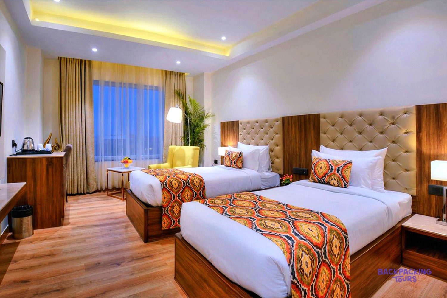 A modern hotel room features two beds with colourful patterned runners, a yellow armchair, and a window.