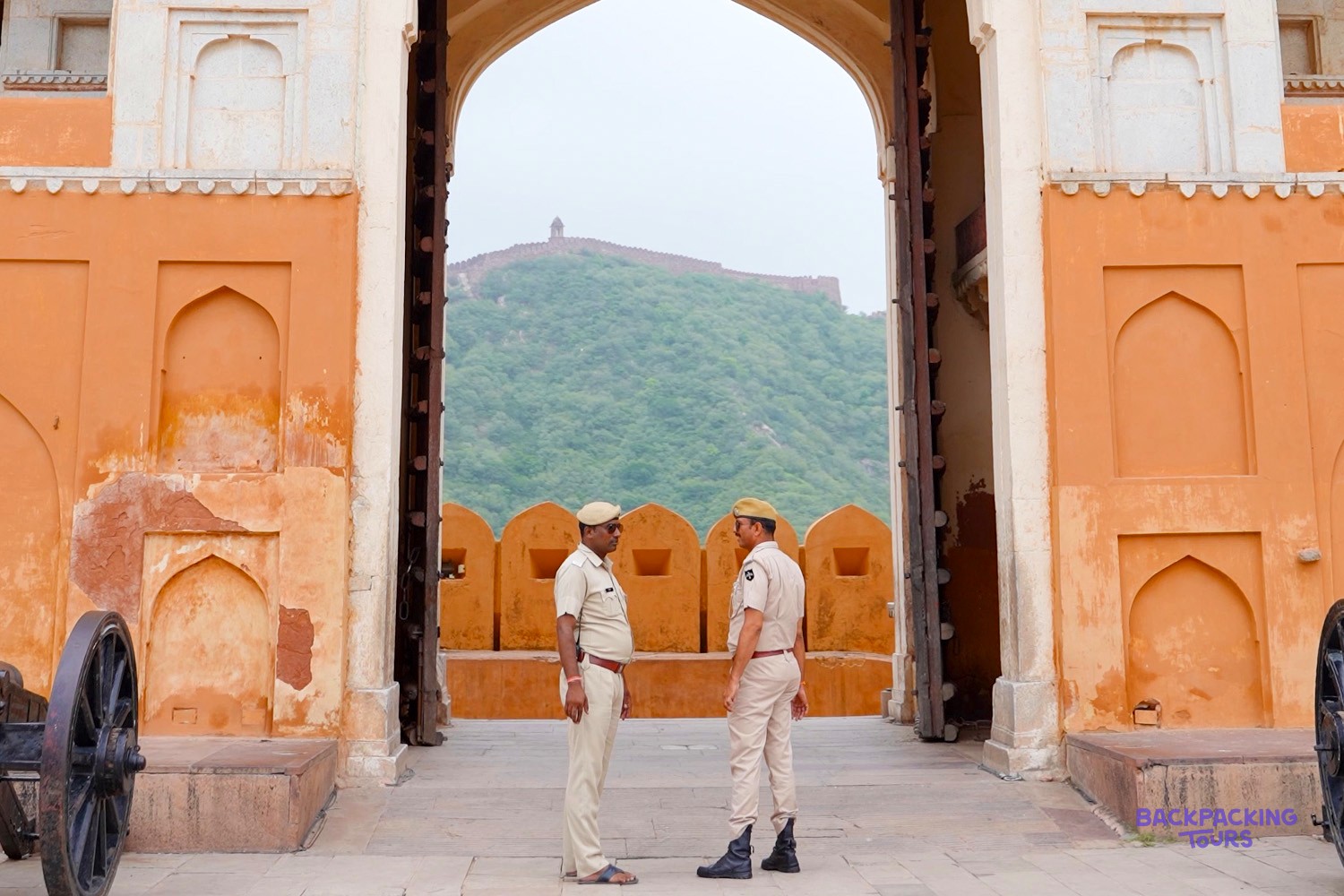 Two uniformed men stand in an arched gateway of an orange fort, with a distant hilltop fort visible.