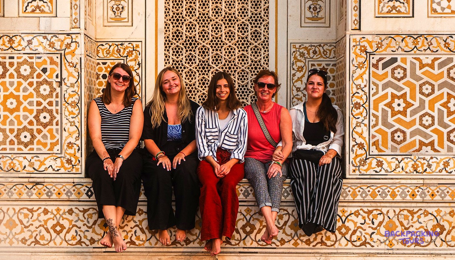 Five women sit barefoot against an ornate wall decorated with yellow and grey geometric patterns.