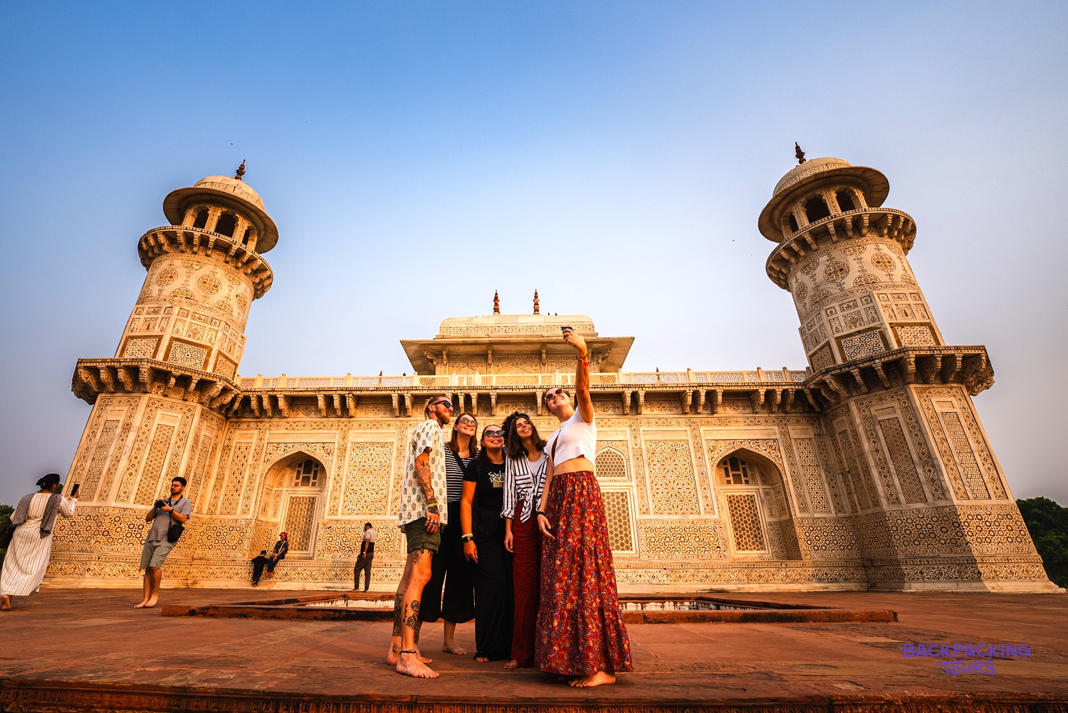 Five young people take a selfie in front of an intricately carved white marble building with two minarets.