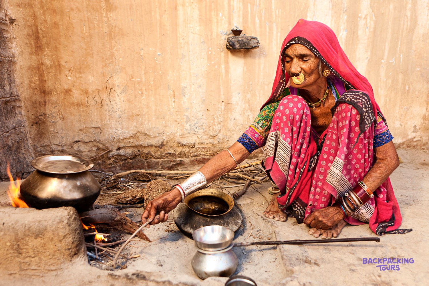 An elderly woman in colourful traditional dress squats, stirring a cooking pot over an open fire.