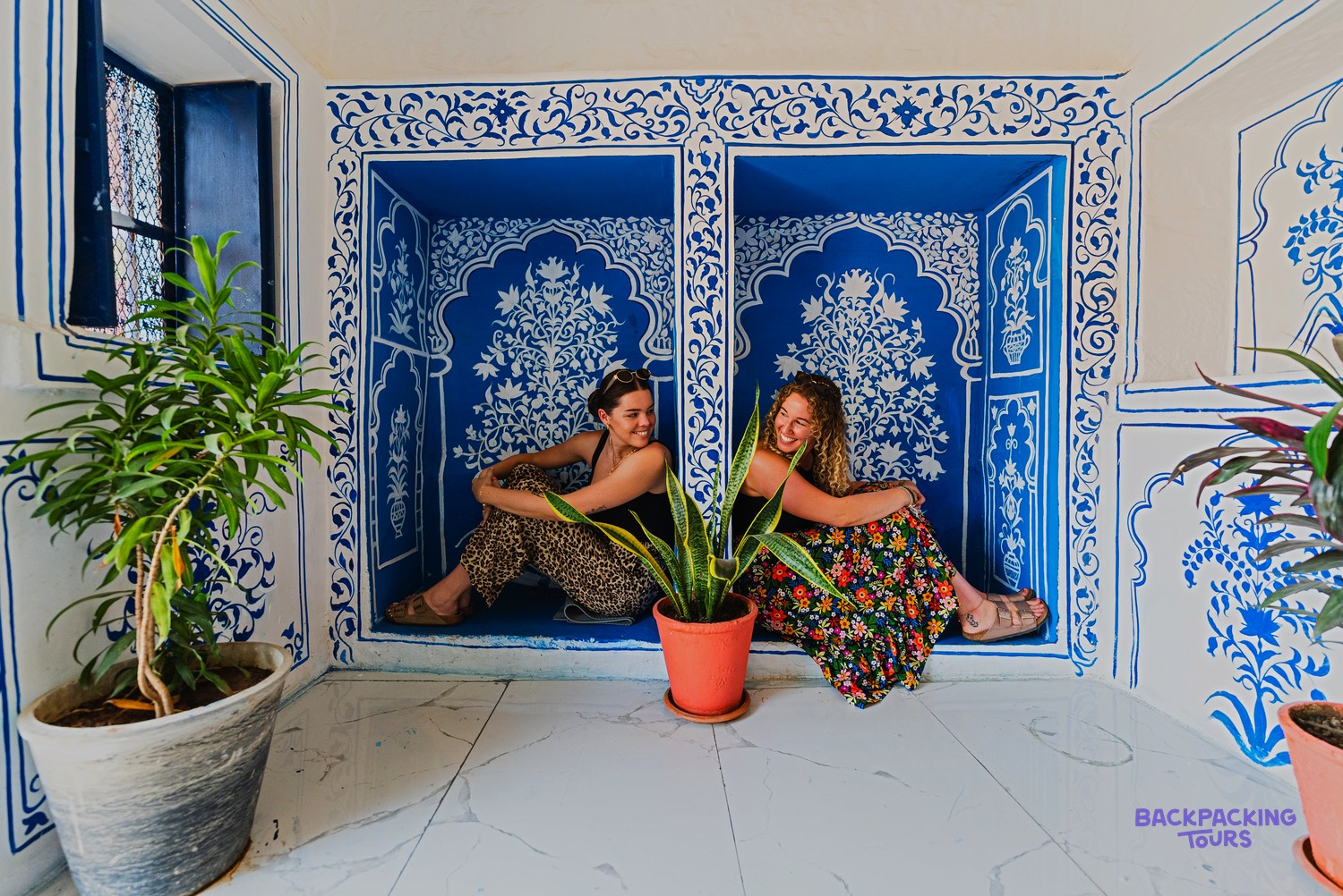 Two women sit in decorative blue alcoves with white patterns, surrounded by potted plants.