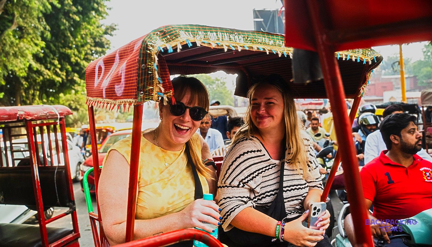 Two women are smiling and laughing while travelling in a red rickshaw on a busy street.