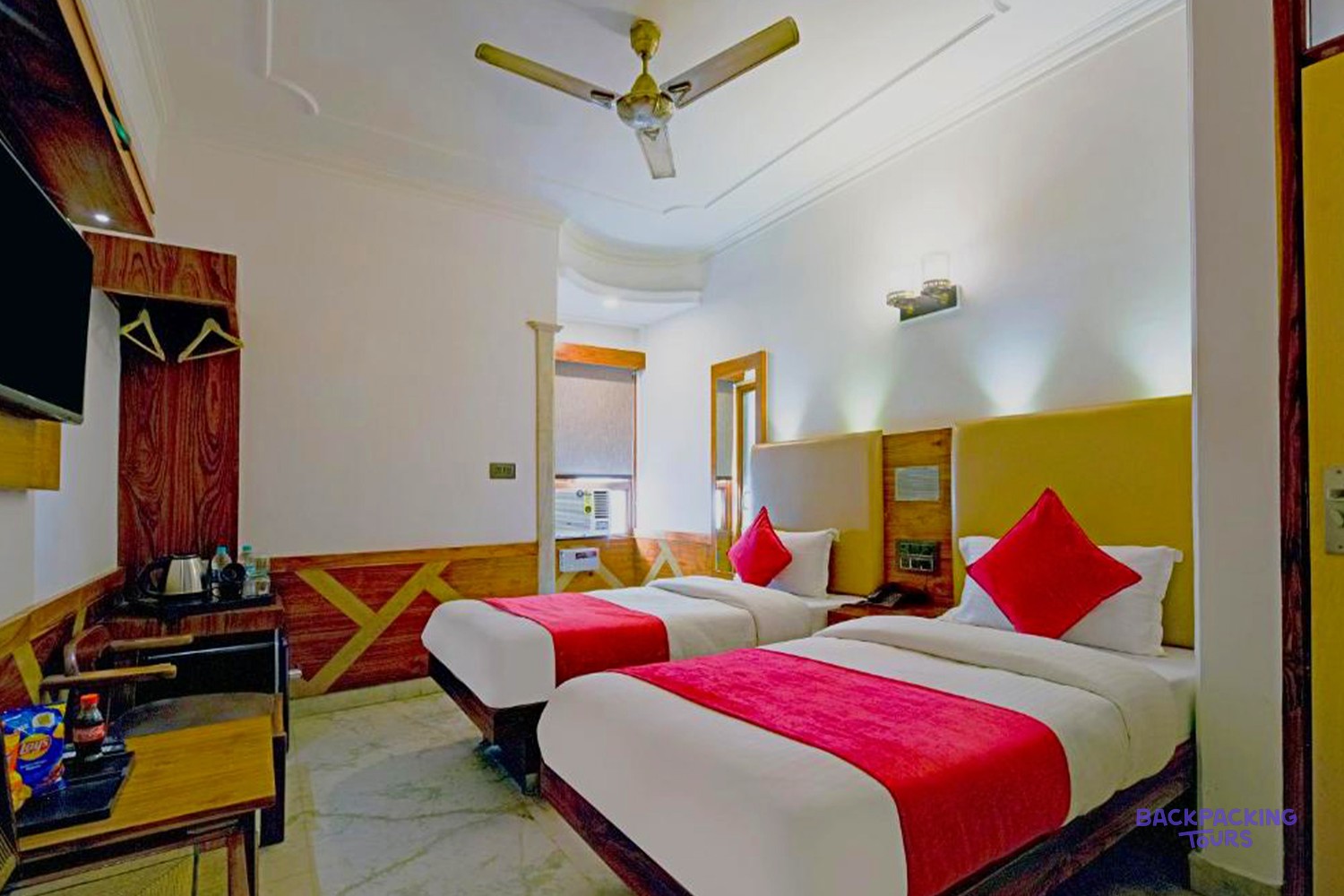 A bright hotel room features two single beds with red throws, a ceiling fan, and a small desk.